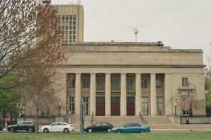 Walker Memorial at MIT, a neoclassical hall with tall columns and inscription dedicated to Francis Amasa Walker, cars parked along the street and bare spring trees in front