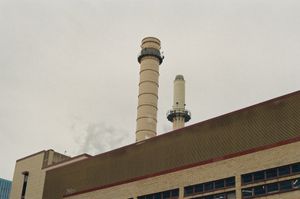 Two tall off-white industrial smokestacks with metal catwalks rising above a brick and corrugated-metal building, faint white smoke against an overcast gray sky
