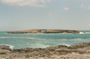 Low flat islet offshore with a natural rock arch framing the ocean beyond, surrounded by turquoise water and white surf under pale blue sky, rocky foreground shore