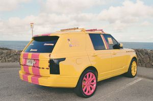 Bright yellow Range Rover with hot pink wheels and racing stripes parked by a stone seawall overlooking the ocean, with Hawaii license plate PRYNX and social media decals on the window