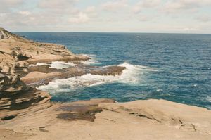 Layered tan and brown volcanic cliffs on Oahu's southeastern shore with white surf breaking over tidal shelves into deep blue Pacific, coastal highway visible on the headland
